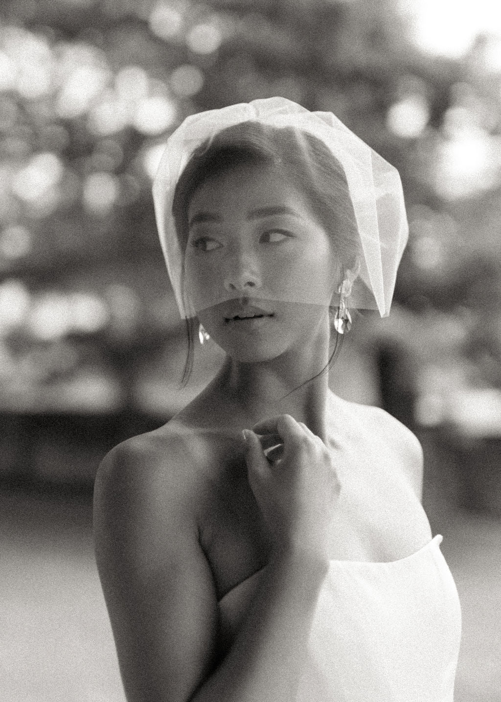Black and white portrait of a woman wearing a sheer white bridal veil and a white strapless dress, with a softly blurred outdoor background.