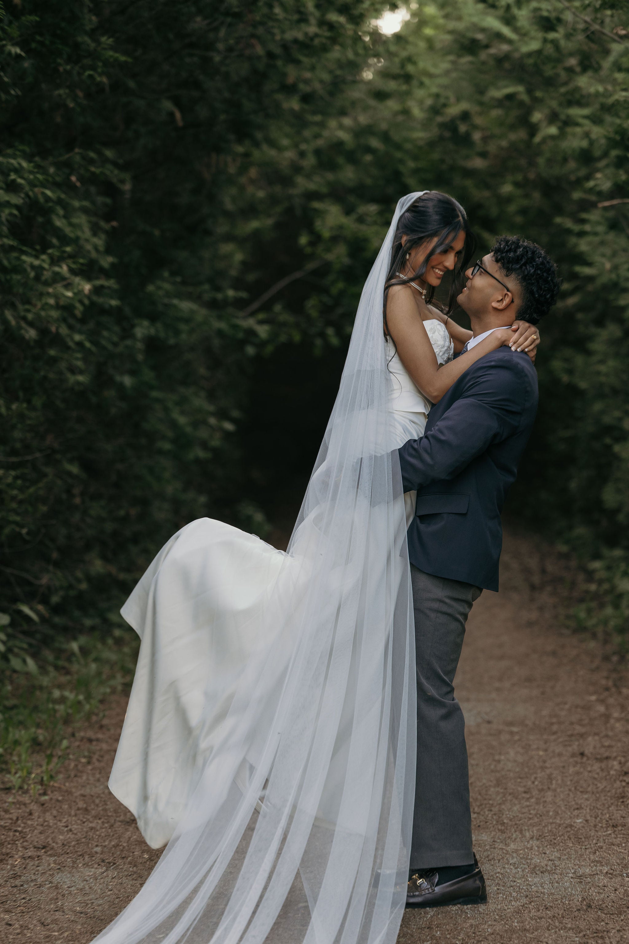 A bride in a white wedding gown with a long sheer veil is being held in a groom's arms on a wooded path. The bride's face is obscured by a rectangular privacy block, and the background consists of dark green trees.