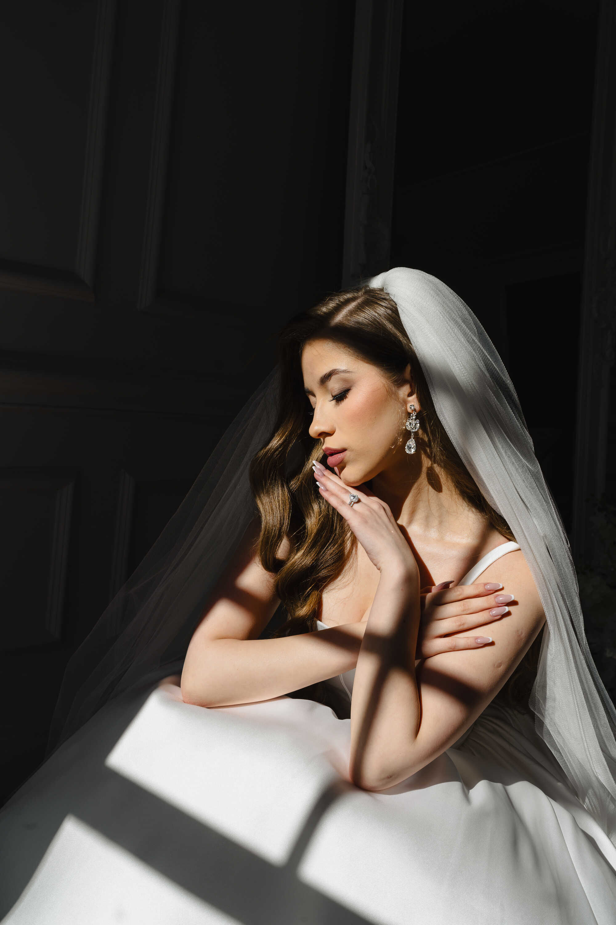Bride in a white wedding dress with a veil, sitting against a dark background.