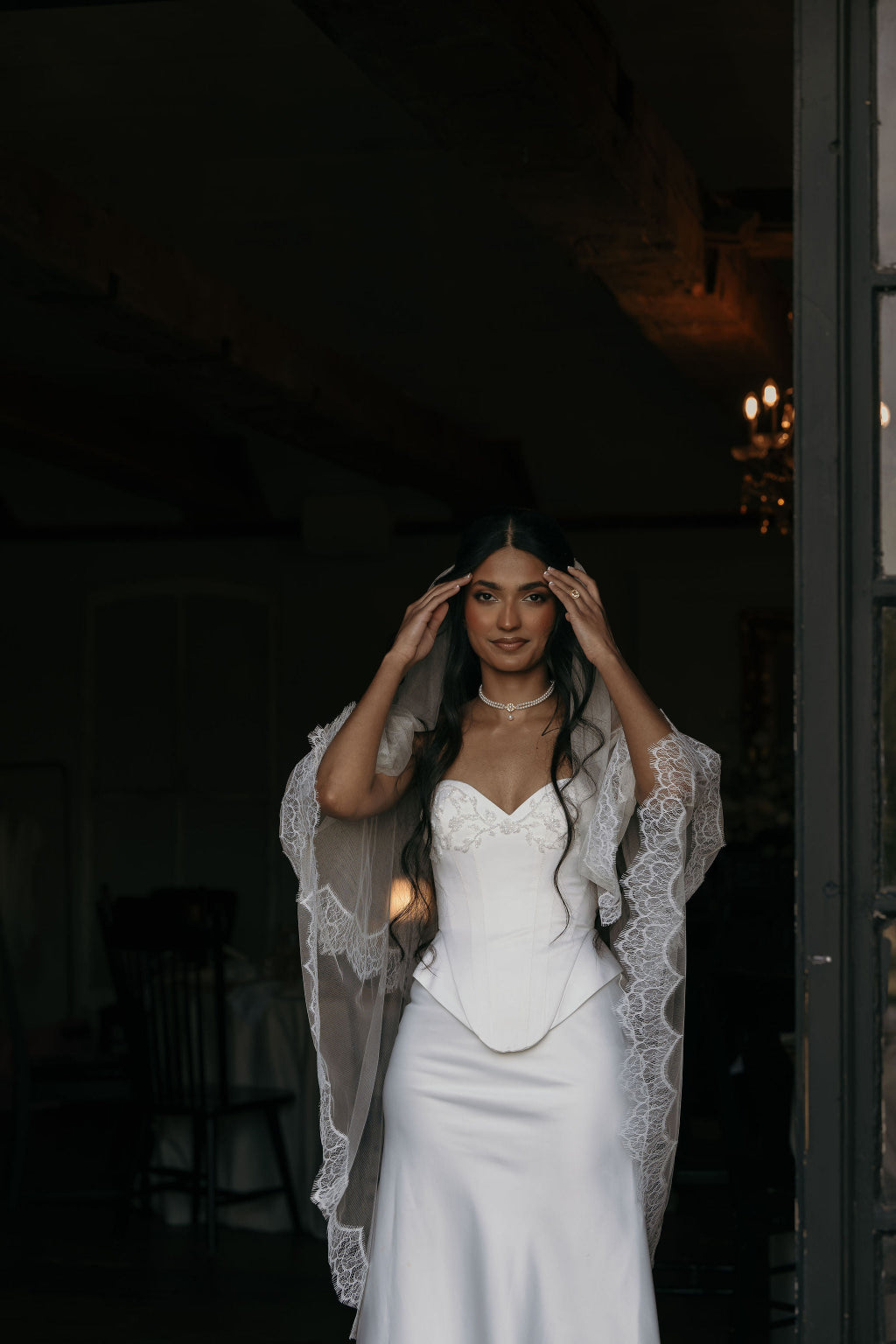 Woman in a white wedding dress with a lace cover-up standing in a dimly lit room.