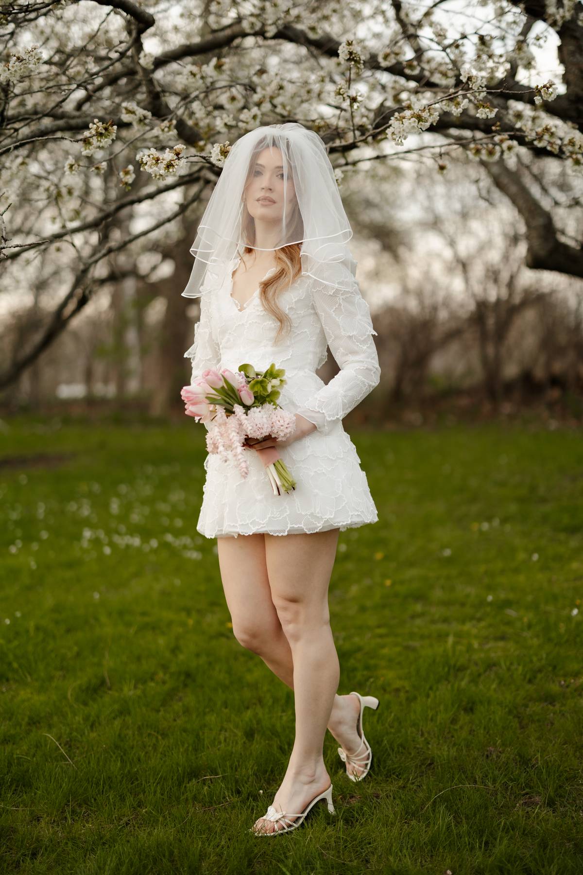 A woman wearing a short white lace bridal dress, veil, and holding a pink bouquet stands on a grassy field under blooming blossom trees.