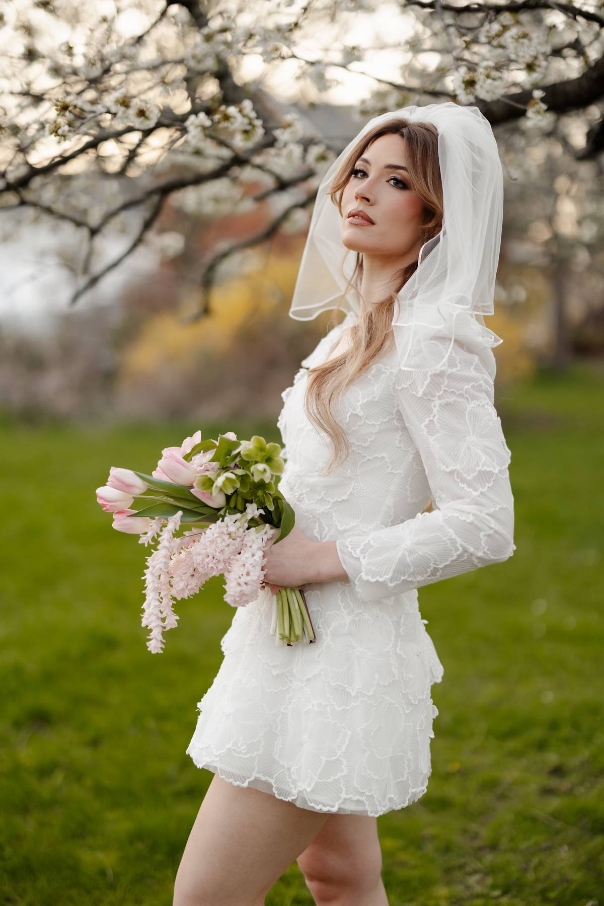 A woman wearing a white lace bridal mini dress with a matching veil stands outdoors on a grassy lawn under blooming trees, holding a bouquet of pink tulips and white flowers.