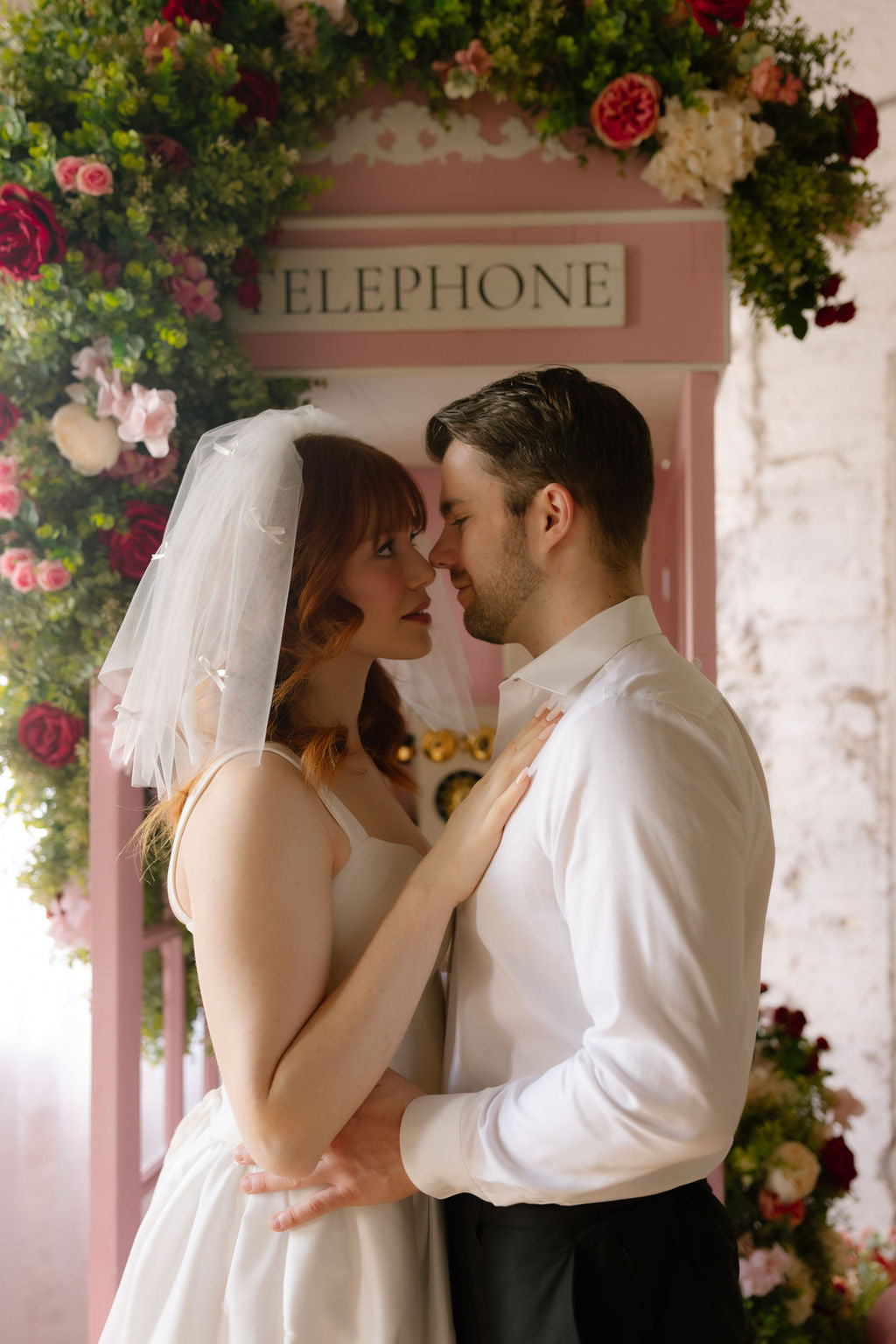 Couple in wedding attire standing in front of a pink telephone booth with floral decorations.
