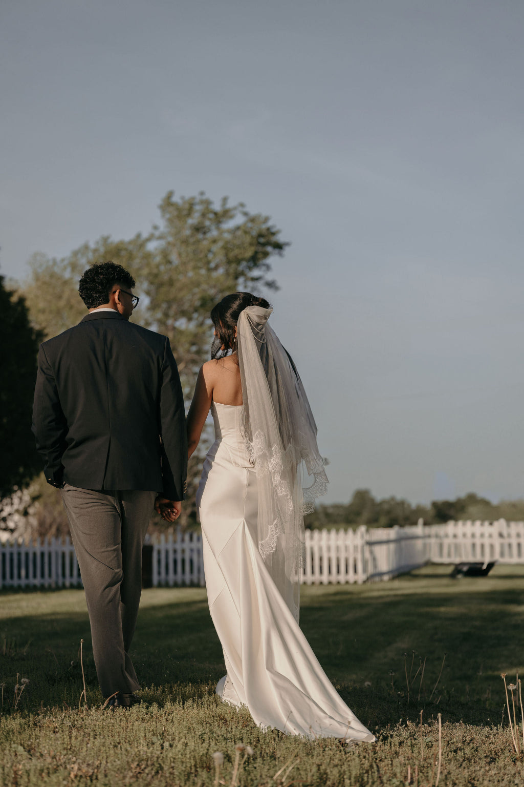 Couple holding hands in a field with a white picket fence and trees in the background.