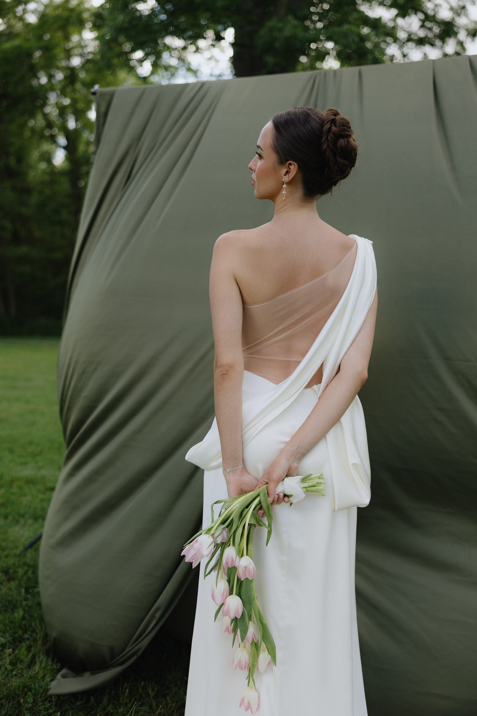 A woman in a white draped one-shoulder gown stands outdoors in front of an olive green fabric backdrop, holding a bouquet of pink tulips.