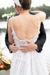 Person in a white wedding dress with an open back and pearl back necklace, held by another person, with a scenic background.
