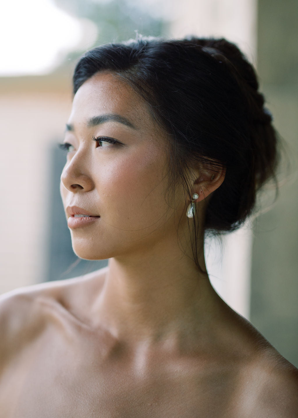 A close-up portrait of a woman in profile wearing a small teardrop-shaped pearl earring, with her dark hair styled in an updo against a softly blurred neutral background.