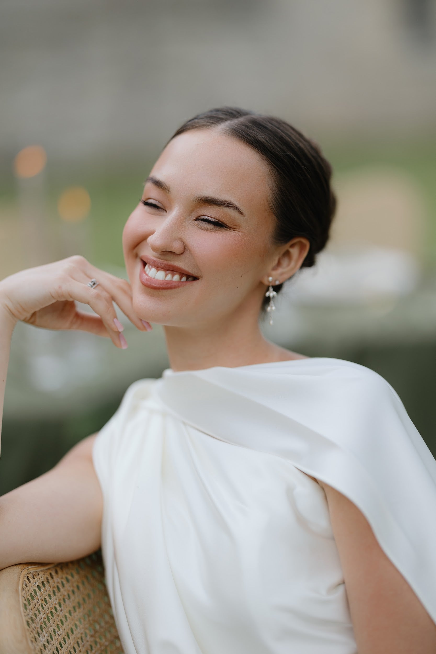 A woman with dark hair pulled back smiles while wearing a white draped top and silver drop earrings, posed in a softly blurred outdoor setting.
