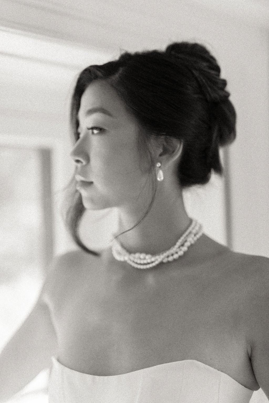 A black and white portrait of a woman with her hair in an elegant updo, wearing a pearl necklace and drop earring, in a softly lit indoor setting.