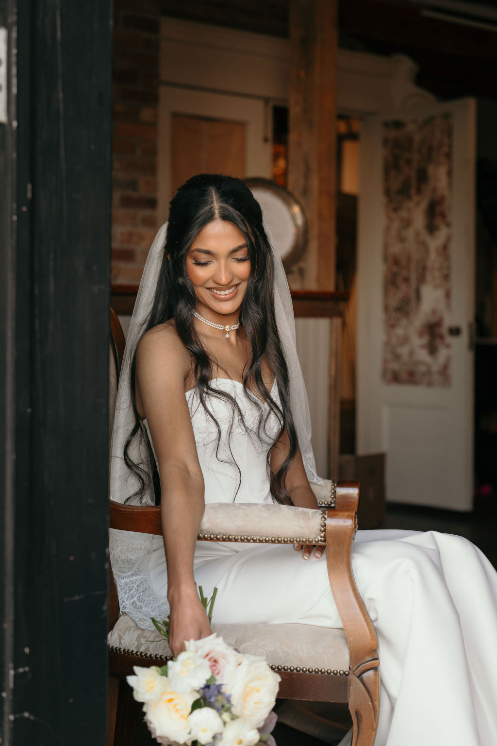 Bride in a white dress with a veil, holding a bouquet, sitting on a chair indoors.