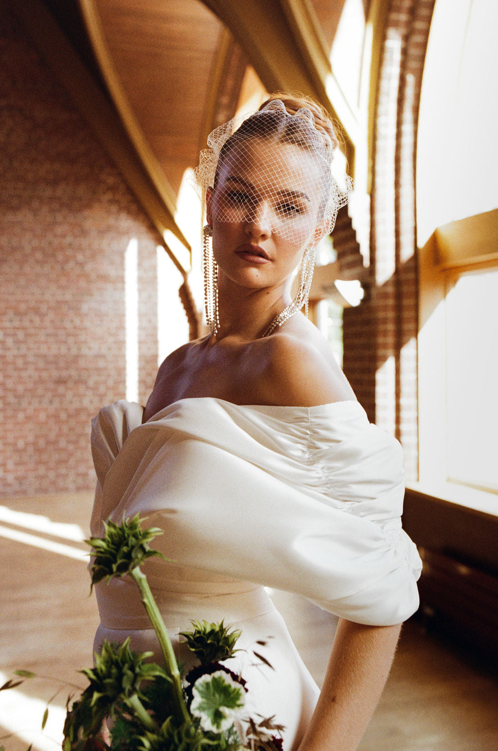 Woman in a white off-shoulder dress with a veil, holding flowers indoors.