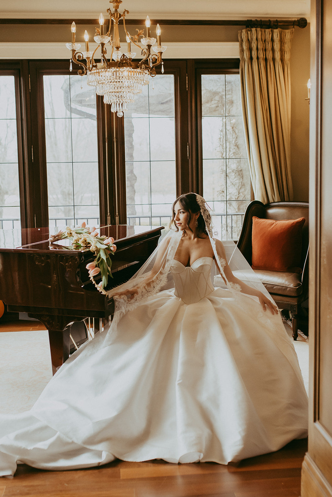 Woman in a wedding dress sitting by a piano in a room with large windows and curtains.