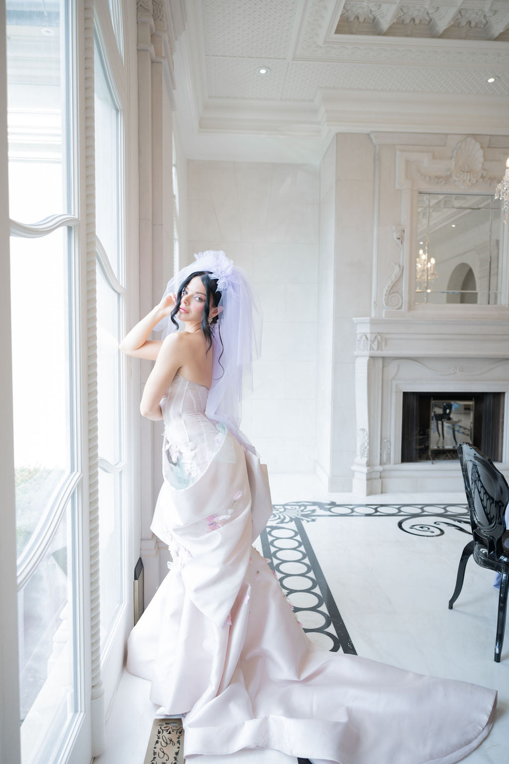 Woman in a white wedding dress standing in a grand room with high ceilings and large windows.