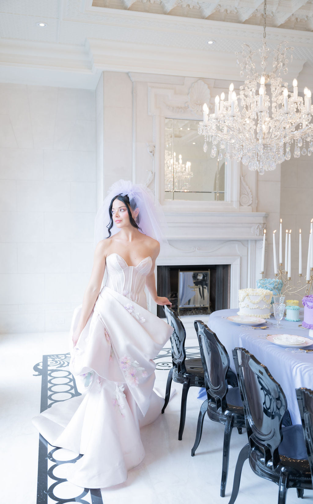 Bride in a white wedding dress standing in a elegant room with chandeliers and a cake on a table.