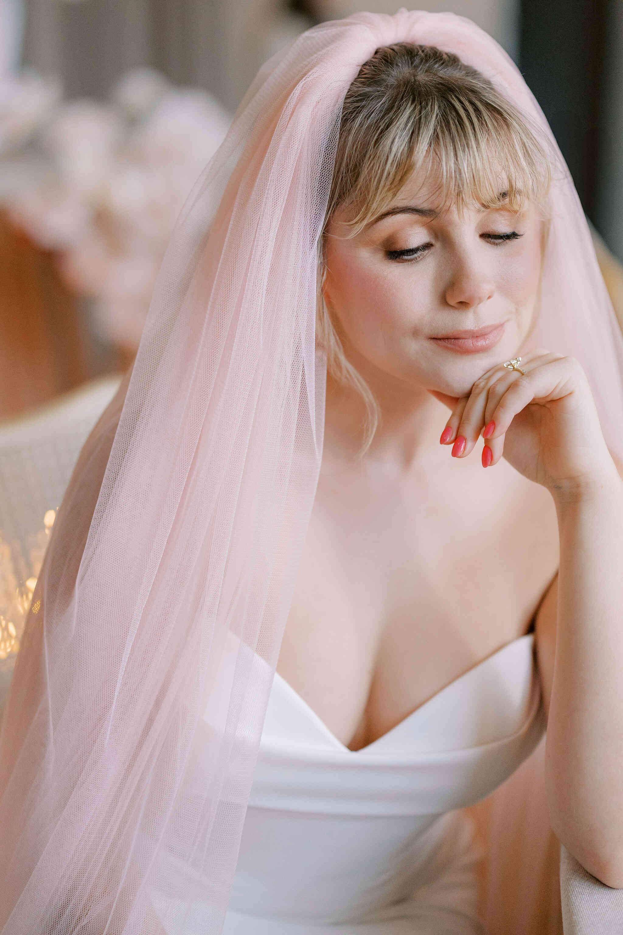 Woman wearing a light pink veil and white dress with red nail polish.