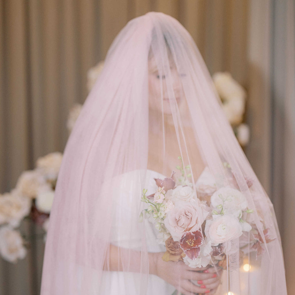 Bride holding a bouquet under a soft pink veil with floral decorations in the background