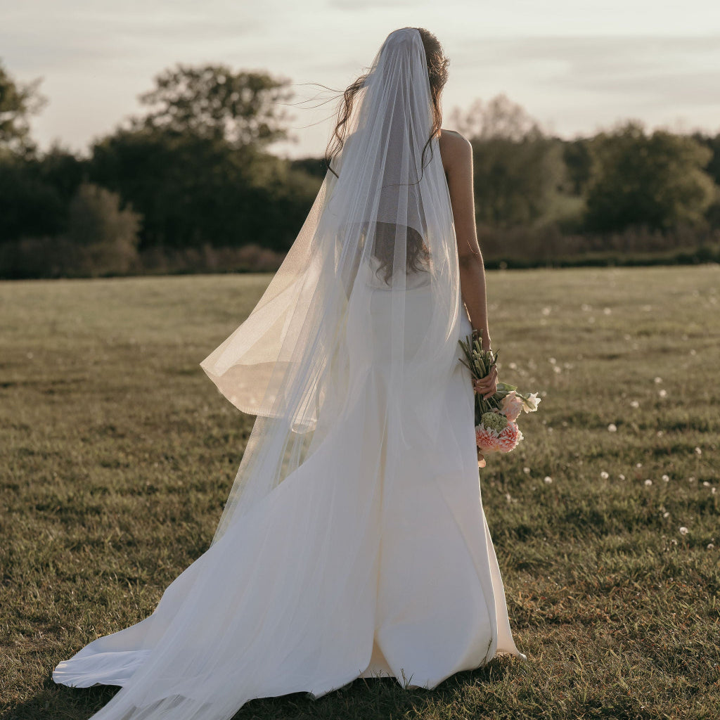 A bride in a flowing white wedding gown and long sheer veil walks away in an open grassy field at sunset, holding a small bouquet.