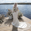 Woman in a white wedding dress standing on a wooden dock by a lake with a scenic background.