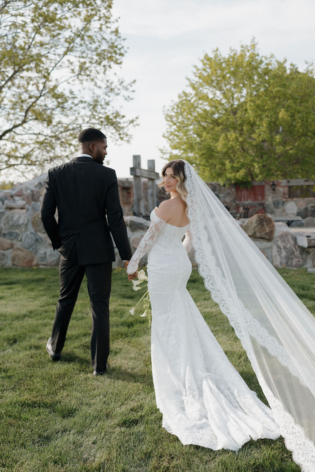 Man and woman in wedding attire standing on grass with a scenic background