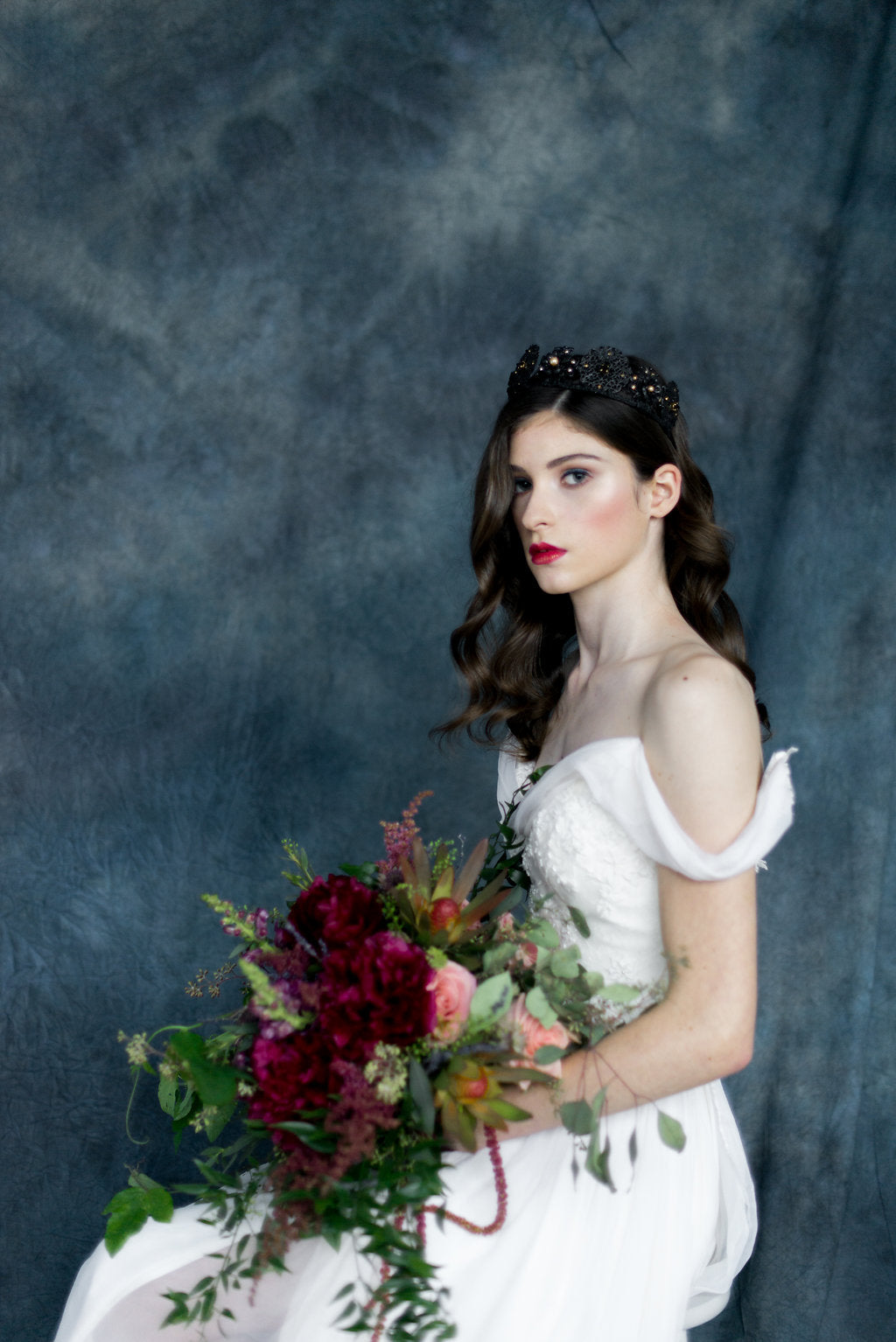 Woman in a white dress holding a bouquet against a dark blue background wearing a black and gold headpiece