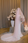 Bride in a white wedding dress with a long pink veil, standing in front of a floral arrangement and candles.
