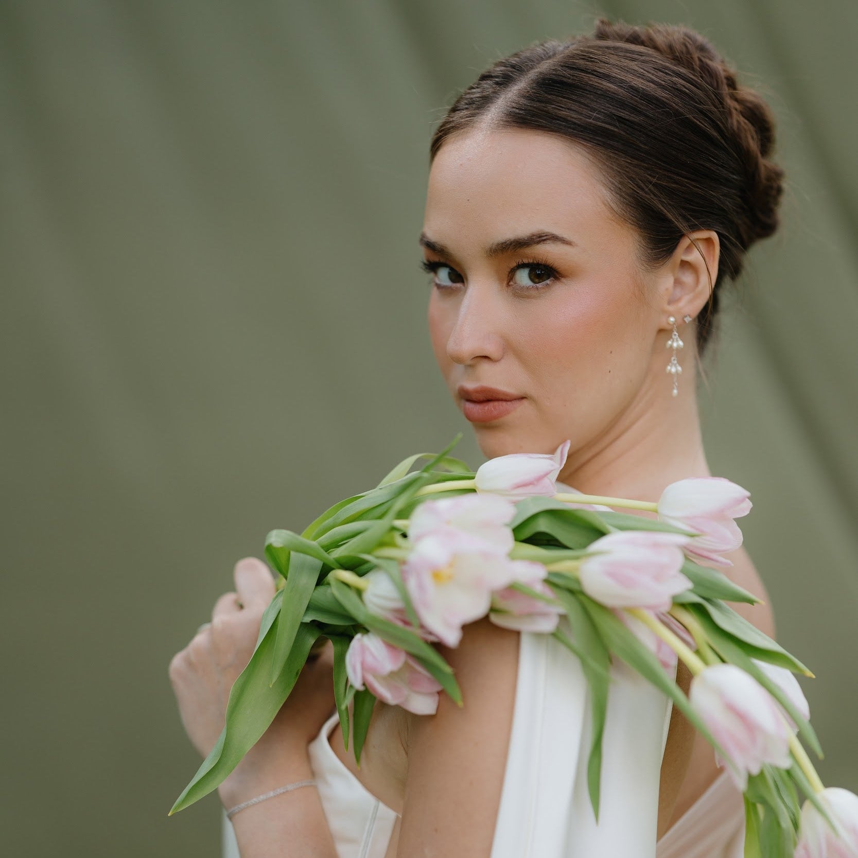 A woman in a white sleeveless dress holds a bouquet of pale pink tulips against a soft olive-green background.