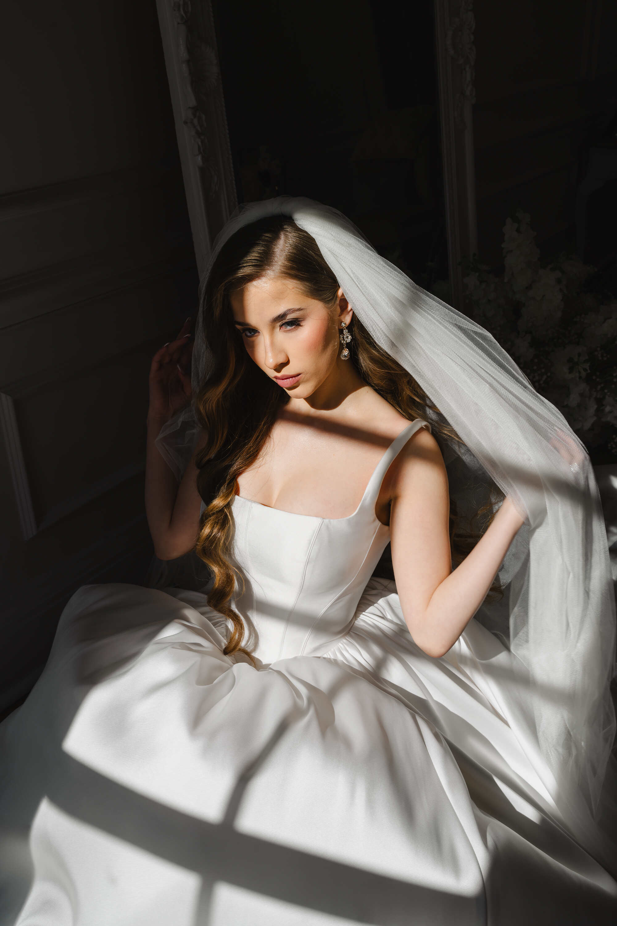 Woman in a white wedding dress with a veil, sitting indoors with soft lighting.