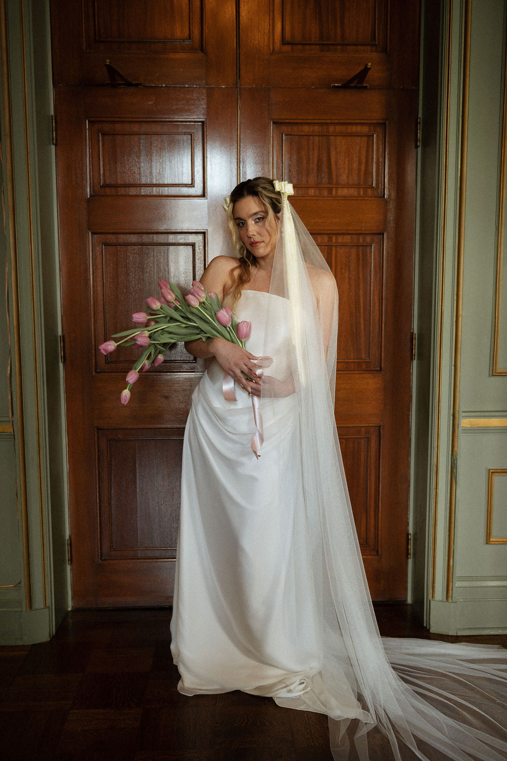 A woman wearing a strapless white wedding gown and long sheer veil stands in front of a large wooden door, holding a bouquet of pink tulips.