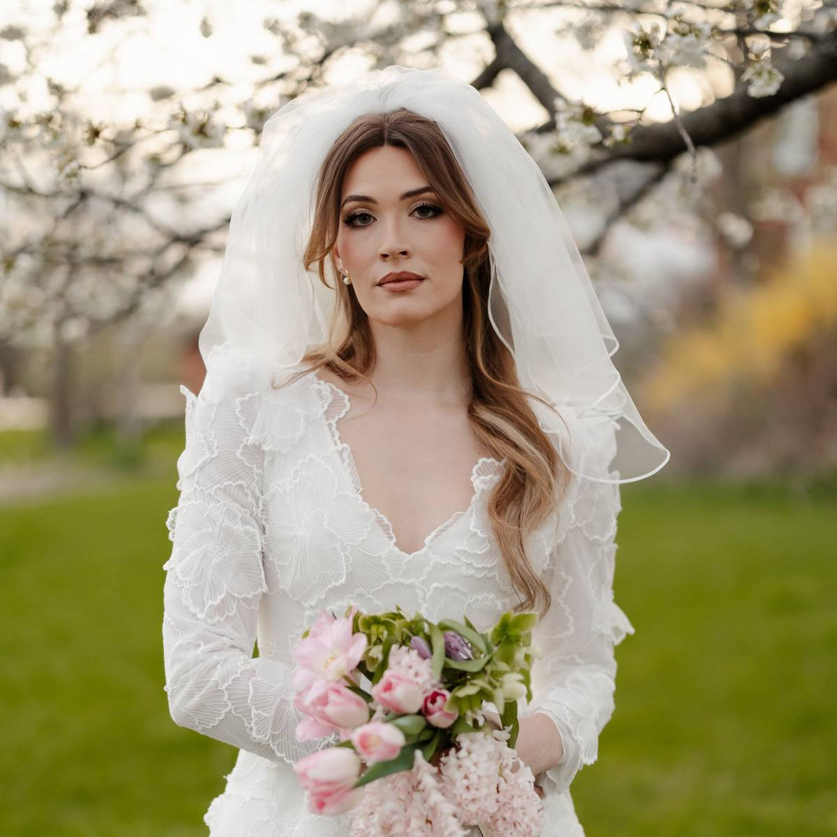A woman wearing a white lace wedding dress with a long veil stands outdoors in a grassy park holding a bouquet of pink flowers.