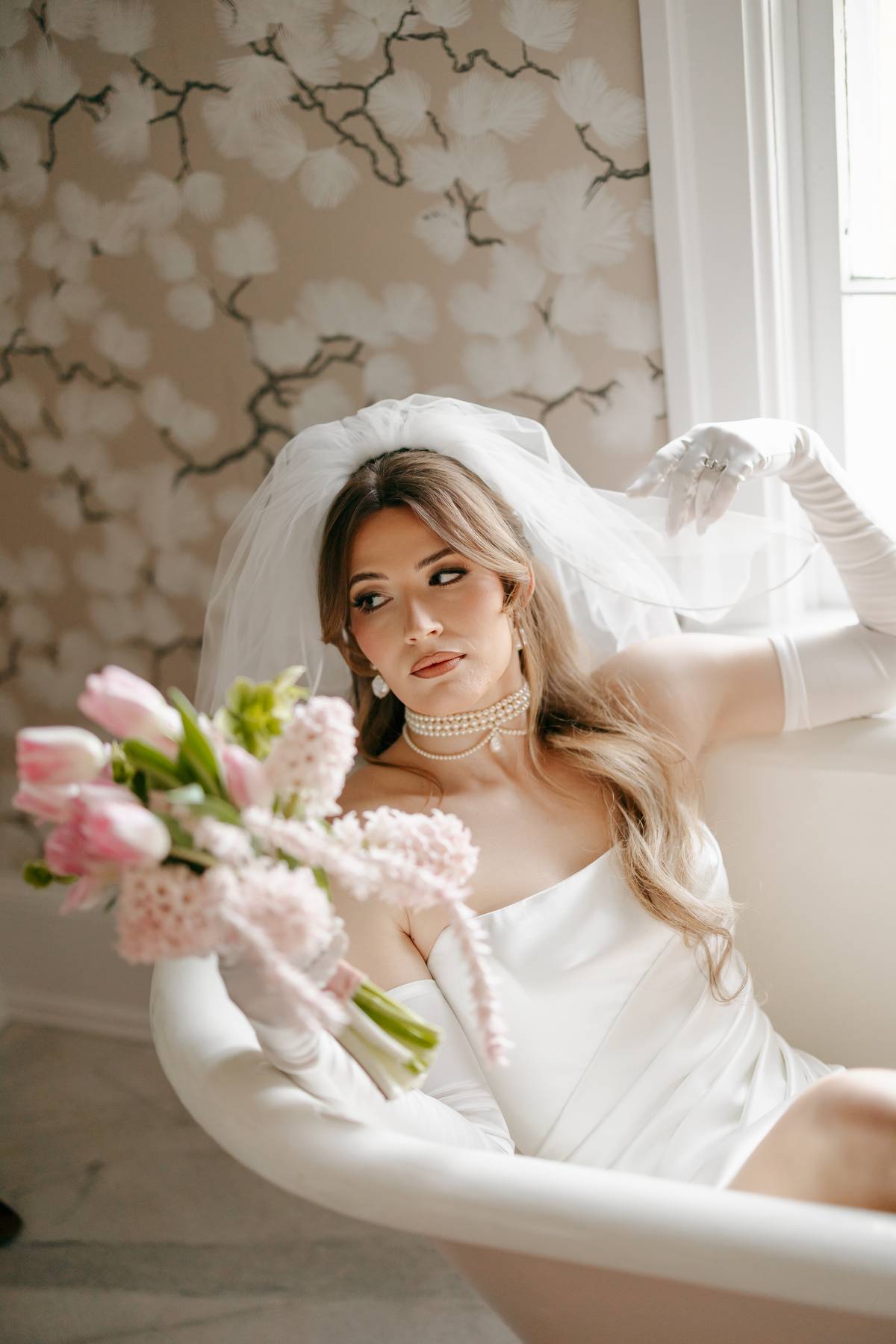 A bride in a white wedding dress, veil, and long white gloves reclines in a white bathtub, holding a bouquet of pink tulips and hydrangeas. She is posed in front of a window with soft light, and the background features a beige floral branch wallpaper.