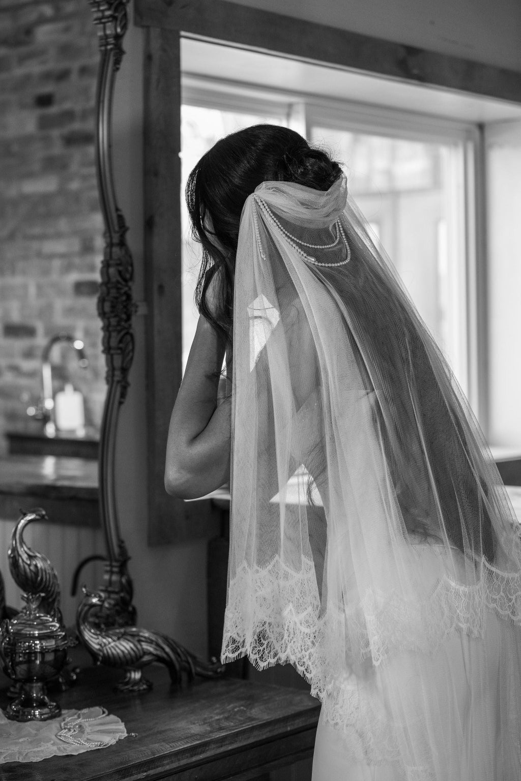 Black and white photo of a bride adjusting her veil in front of a mirror.