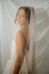 Woman wearing a white wedding dress with a sheer veil against a white curtain background