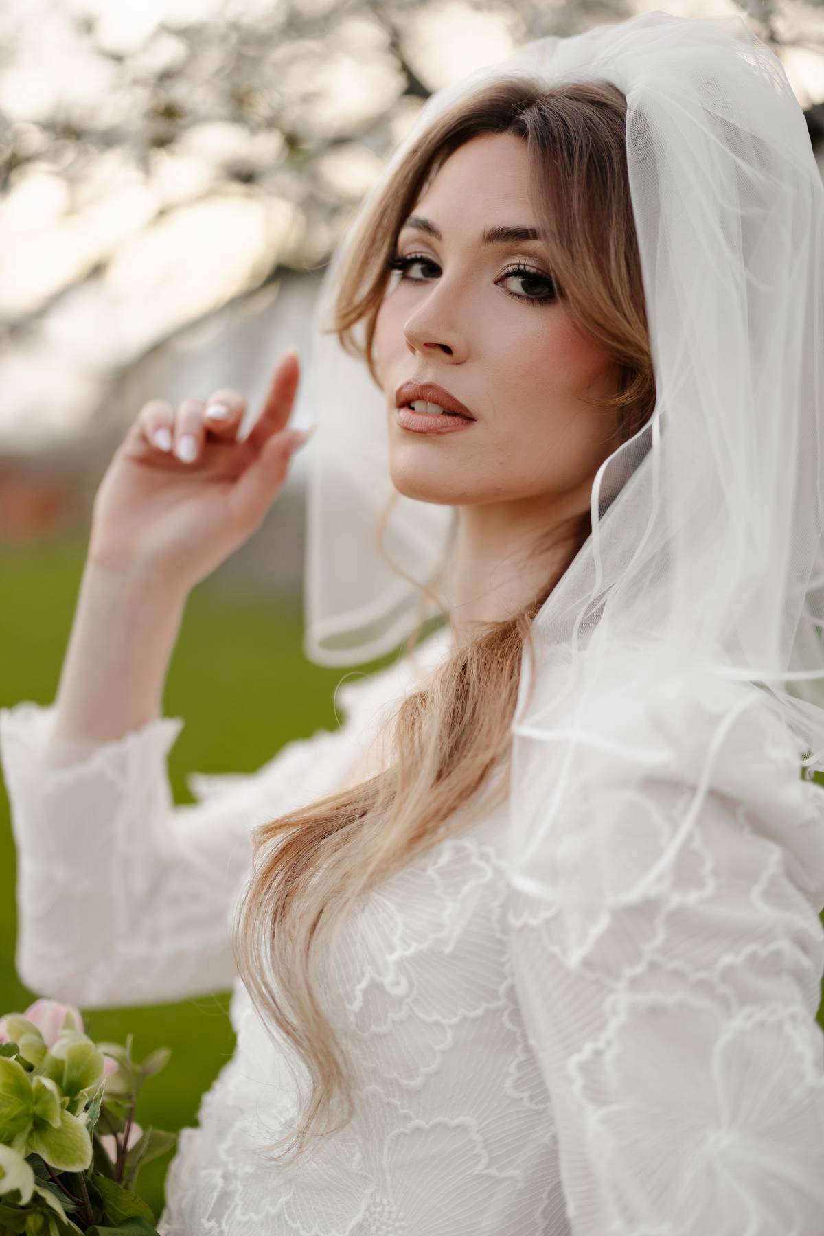 A bride in a white lace wedding dress and matching veil, posing outdoors with a soft-focus background of green grass and trees.