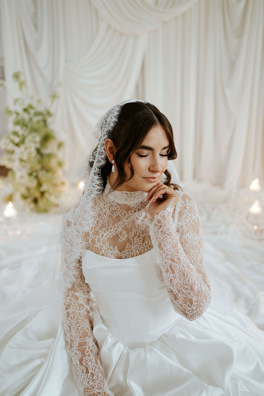 A woman wearing a white wedding dress with long sheer lace sleeves and a matching lace veil, seated in a softly lit bridal setting with draped white fabric and candles in the background.