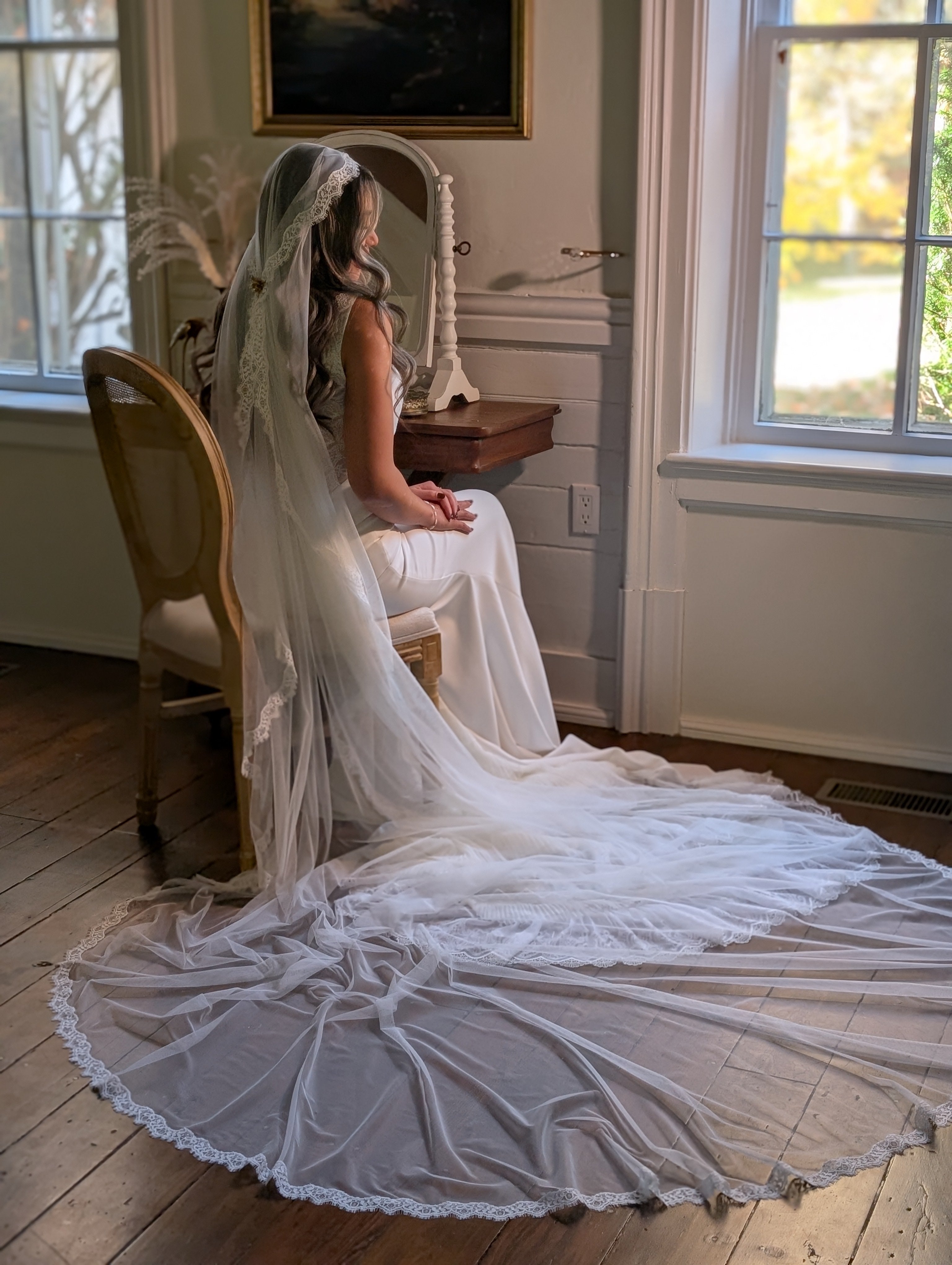 Woman in a white wedding dress with a long veil sitting on a chair in a room with large windows.