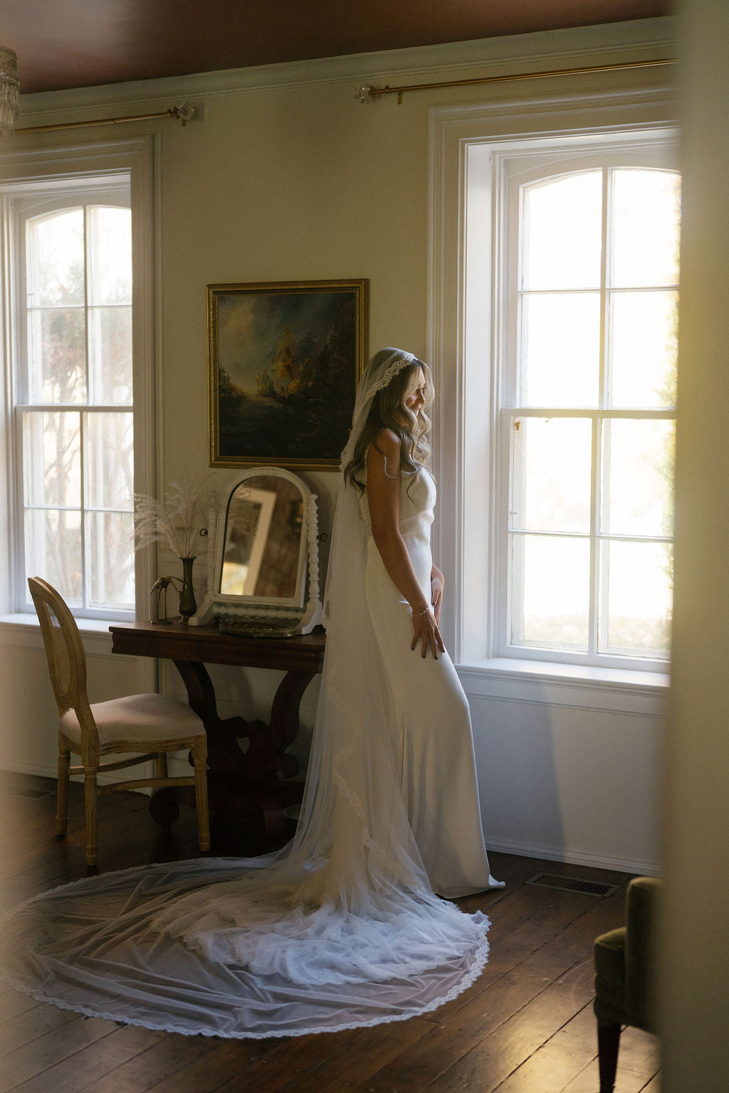 Woman in a white wedding dress standing by a window in a room with a painting on the wall.
