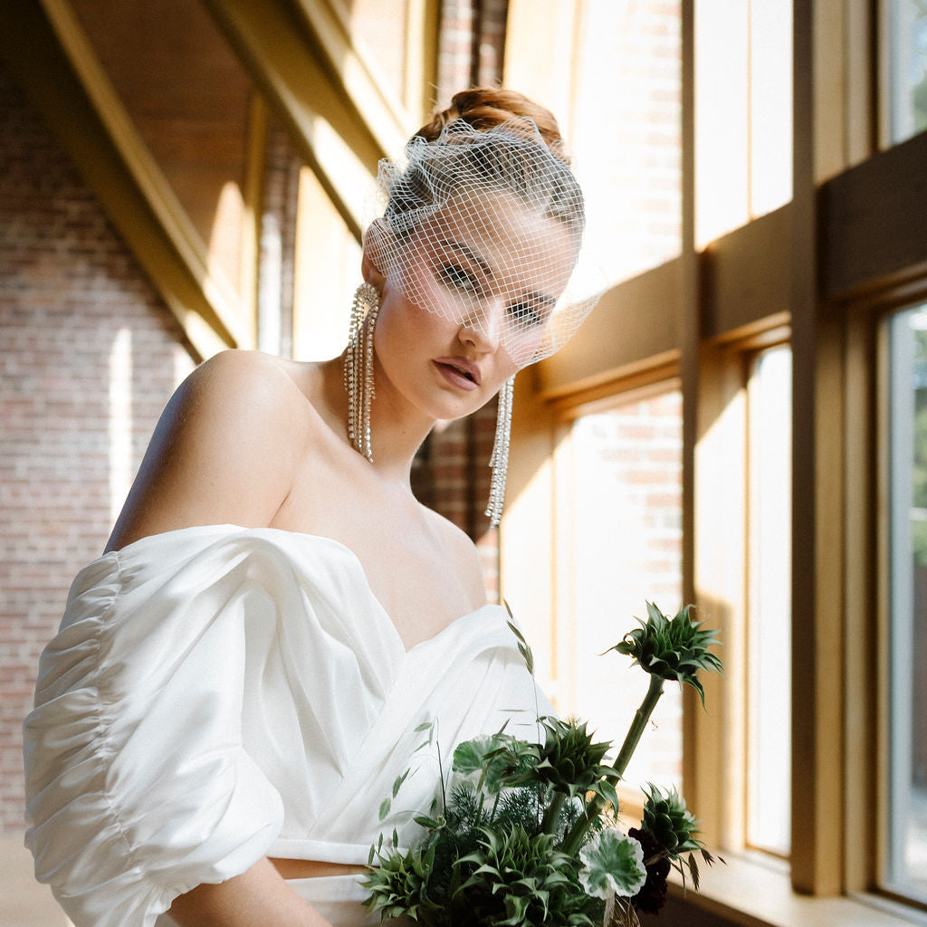 Bride in a white wedding dress wearing a birdcage veil holding a bouquet of green flowers indoors.