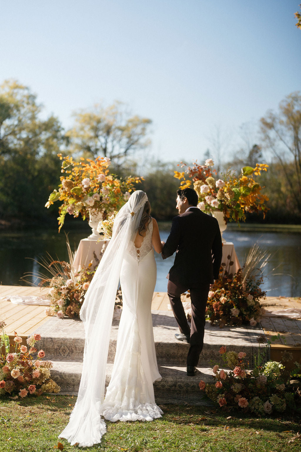 Wedding couple standing on steps by a lake with floral arrangements.