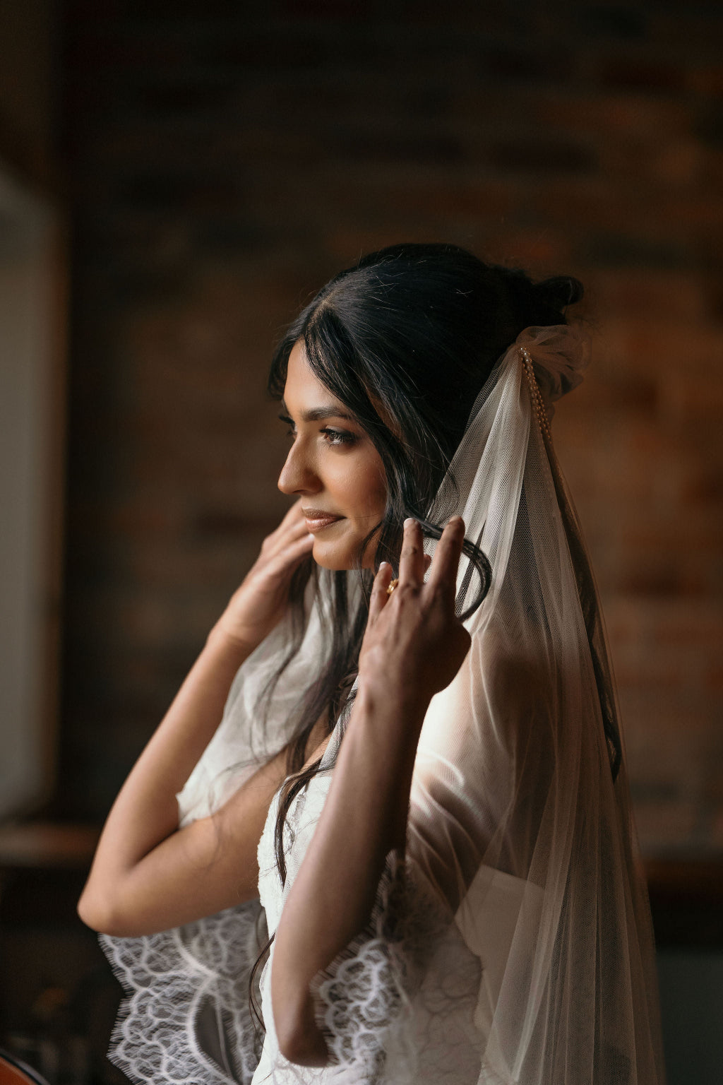 Woman adjusting her veil against a rustic brick wall.