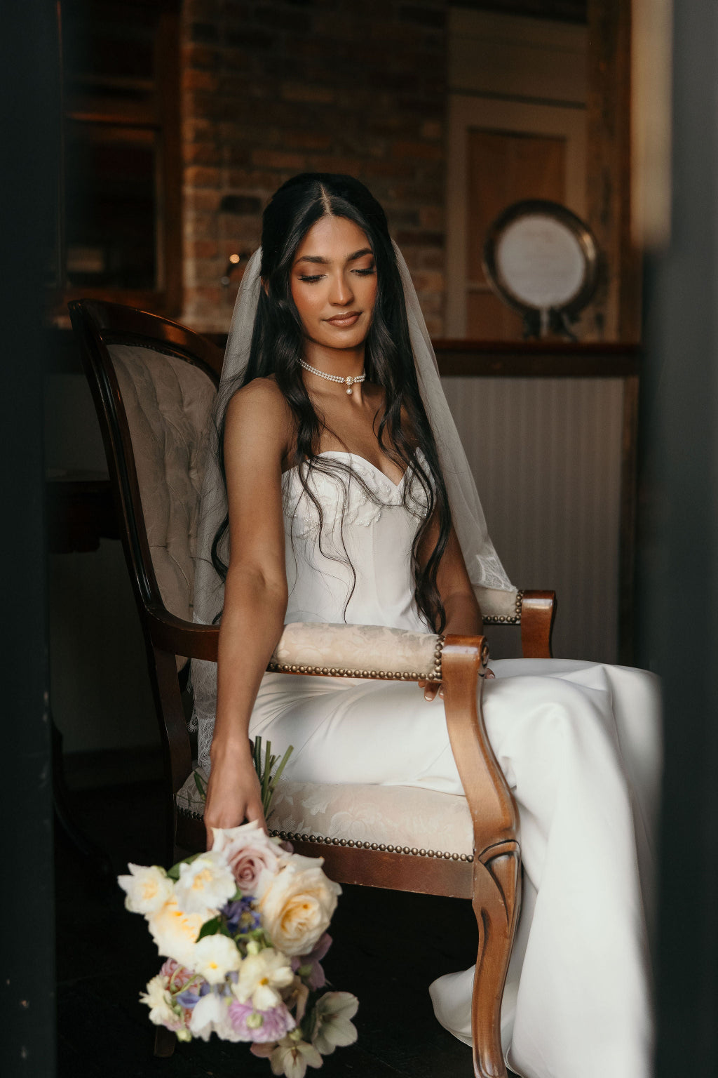 Bride in a white wedding dress holding a bouquet, sitting on a wooden chair.