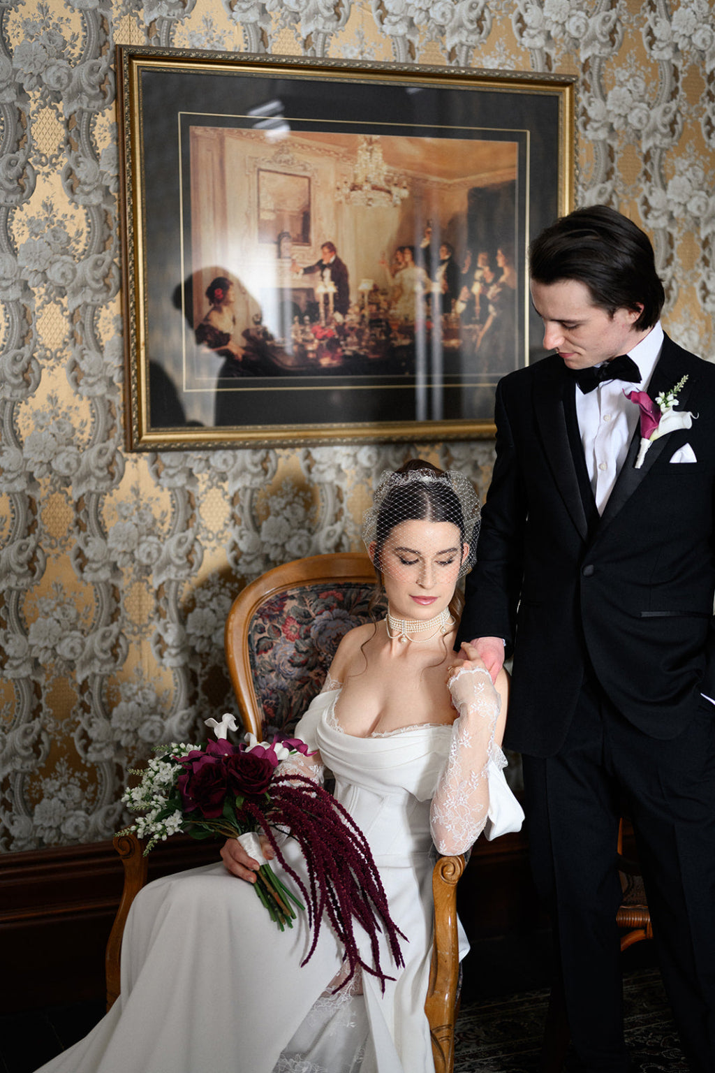 Man and woman in formal attire sitting together with a decorative wall and framed picture in the background.