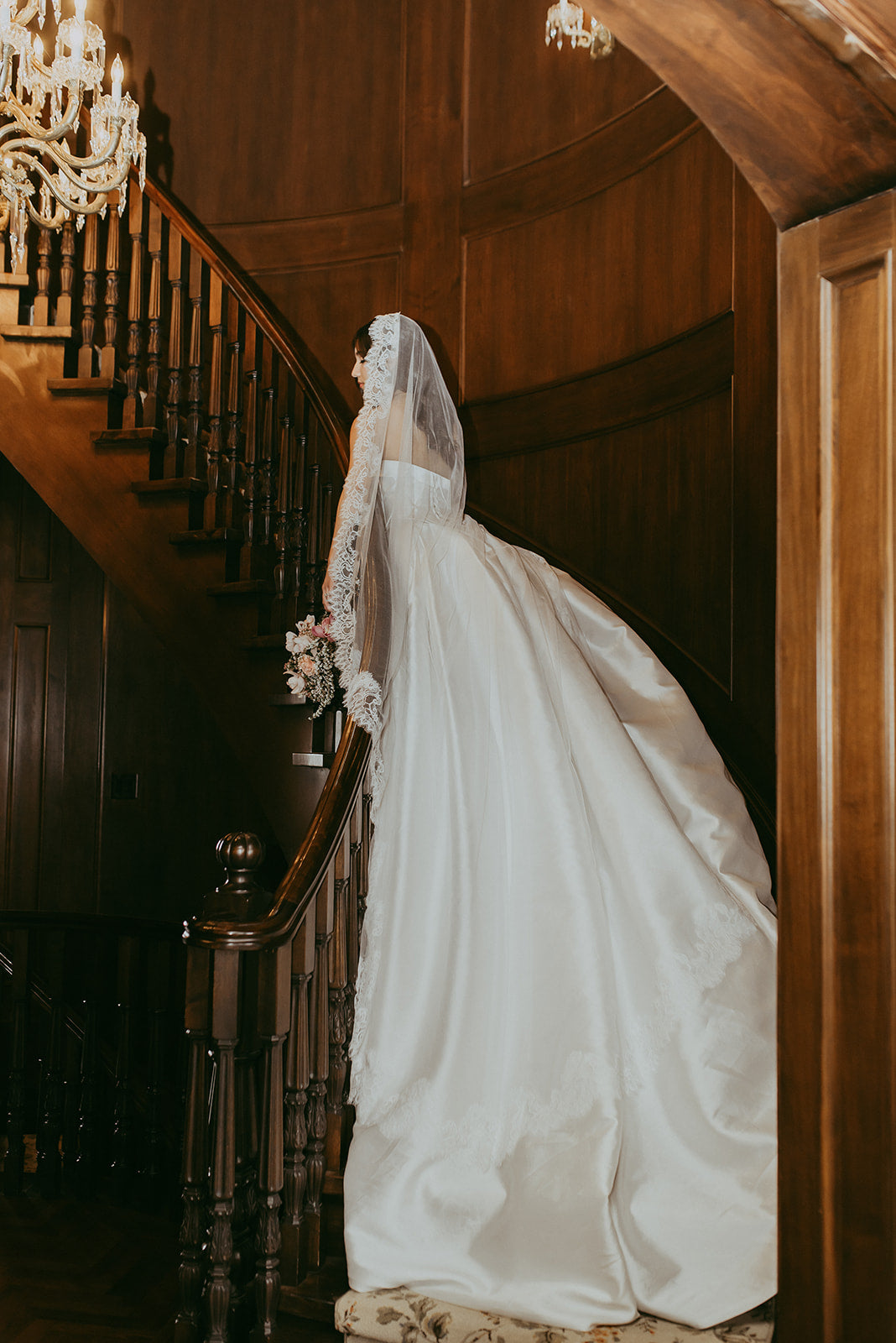 Bride in a white wedding dress standing on a staircase with a chandelier above.