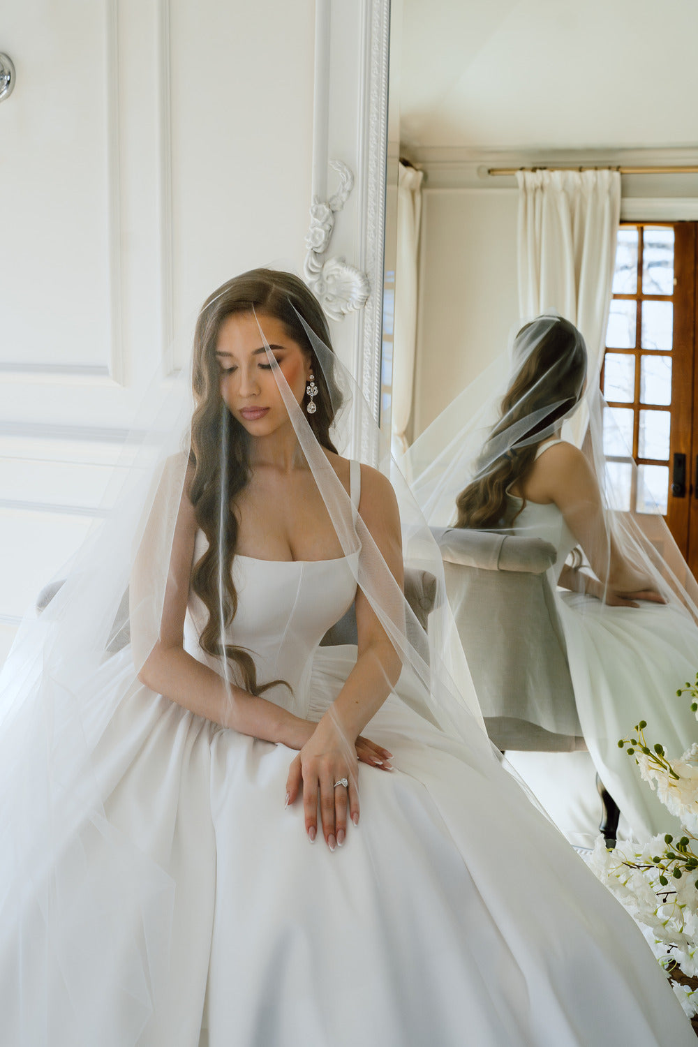 Woman in a white wedding dress sitting on a chair with a veil draped over her head.