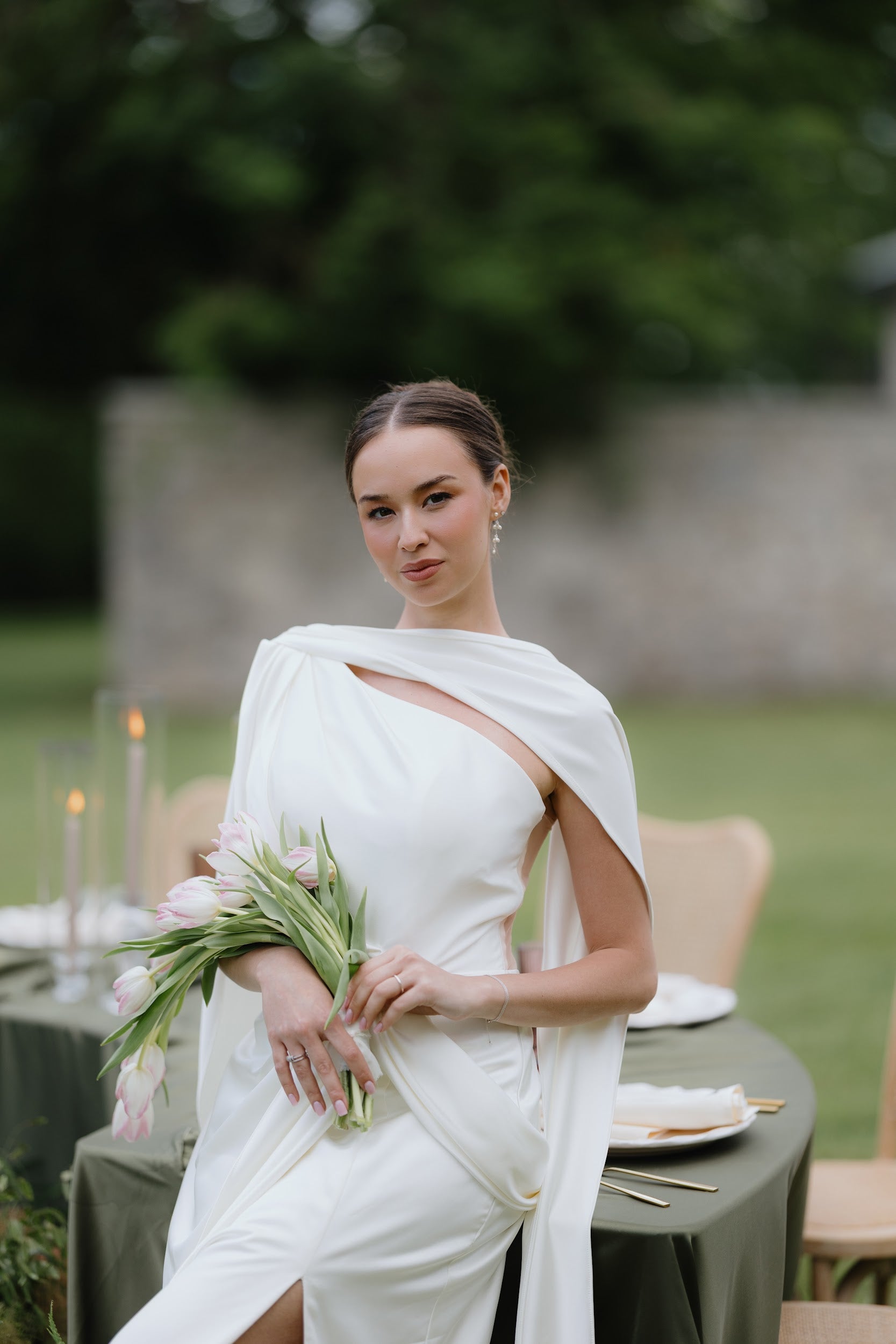 A woman seated outdoors at a table wearing a draped off-white dress and holding a bouquet of pale pink tulips. The background shows a blurred green lawn and stone wall.