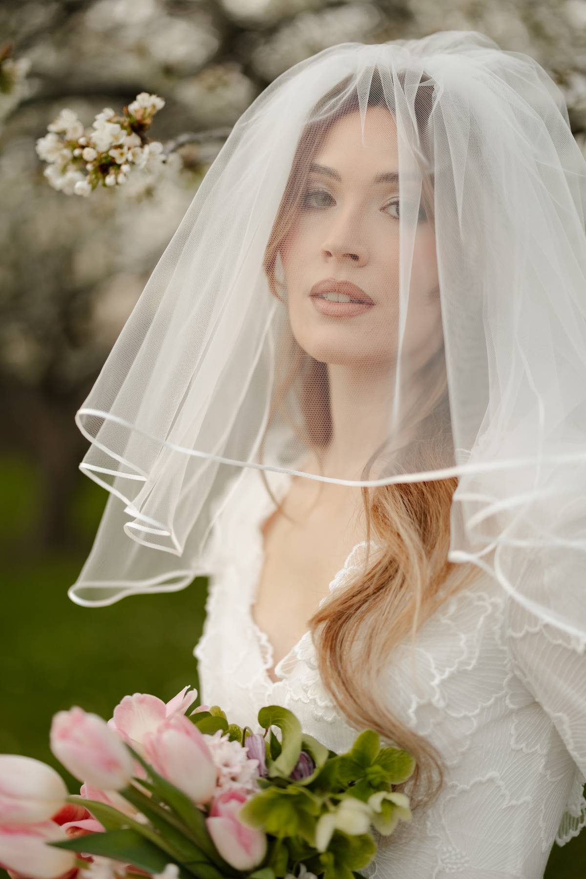 A bride wearing a long white tulle bridal veil and white lace wedding dress, holding a bouquet of pink tulips outdoors among blooming trees.