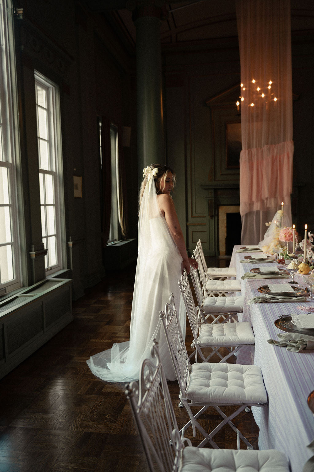 A bride in a white wedding gown and veil stands beside a long wedding table set with white chairs in a dimly lit historic room with tall windows and warm hanging lights.