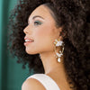 Golden bridal teardrop earrings with pearl studs. Large brass leaves and clusters of clay flowers at the top of the teardrop and bottom. Sprigs of crystals and rhinestones surround the flower clusters. Finished with a larger baroque style pearl. handmade in Toronto canada by Blair Nadeau Bridal Adornments